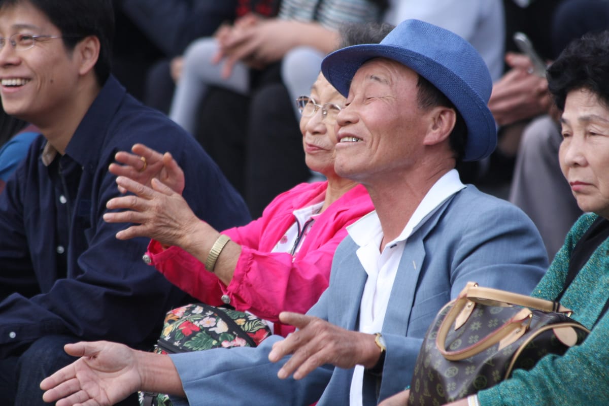 Old Korean man in hat and old Korean woman in pink jacket smiling in audience