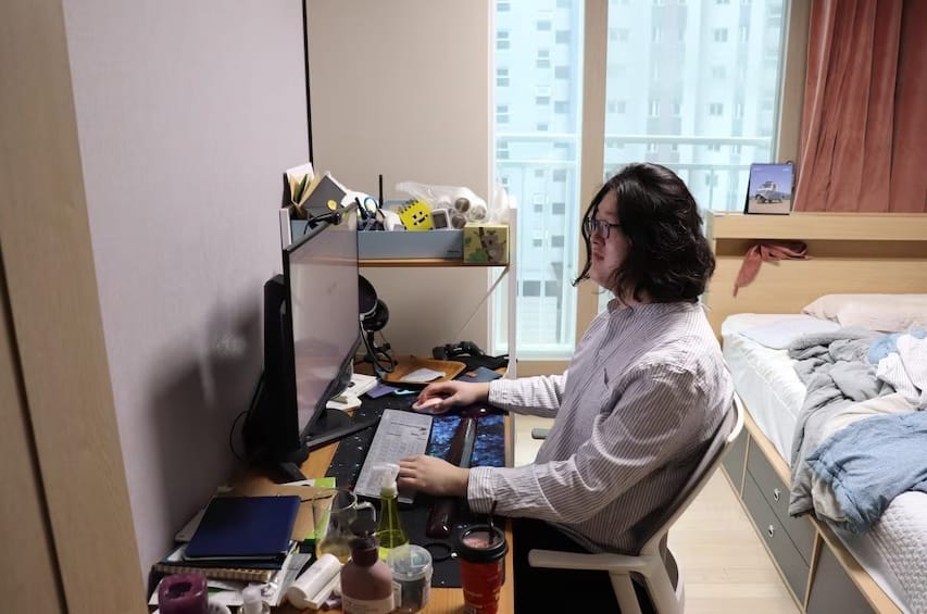 Young man seated at his computer in a bedroom with an unmade bed with highrise view out the window